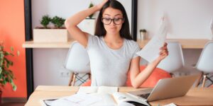 Woman holding paperwork and scratching her head while working at a laptop, representing the challenge of keeping records for a limited company.