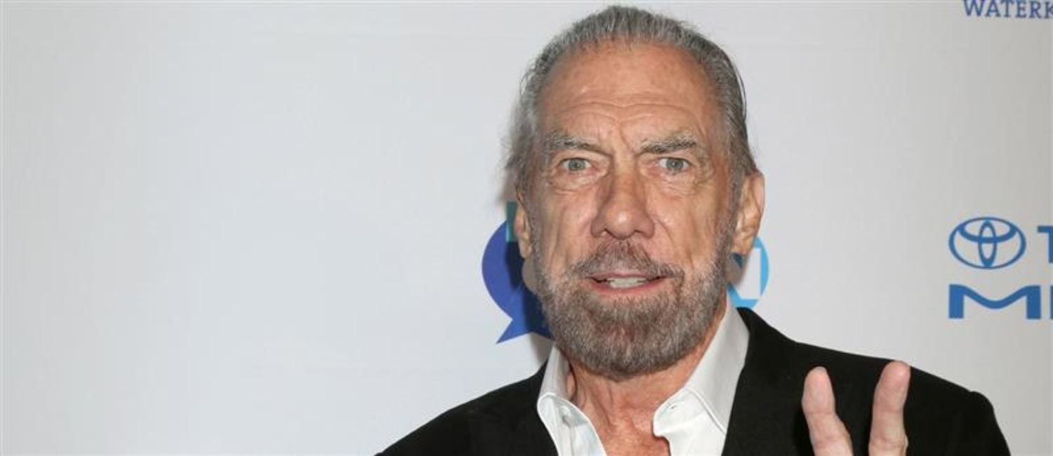 Джон Пол ДеДеория в черном пиджаке поверх белой рубашки, позирующий на белом фоне с логотипами, делает жест примирения рукой.