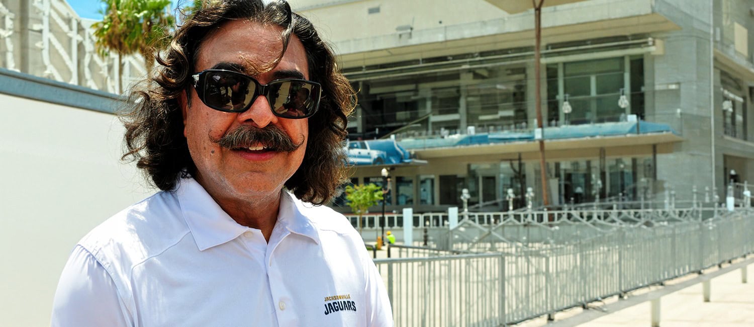 Shahid Khan wearing sunglasses and a white polo shirt stands outdoors, smiling in front of a building with modern architecture.
