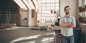 A person stands confidently in a bright workshop filled with plants and art supplies. Large windows allow natural light to illuminate the space.