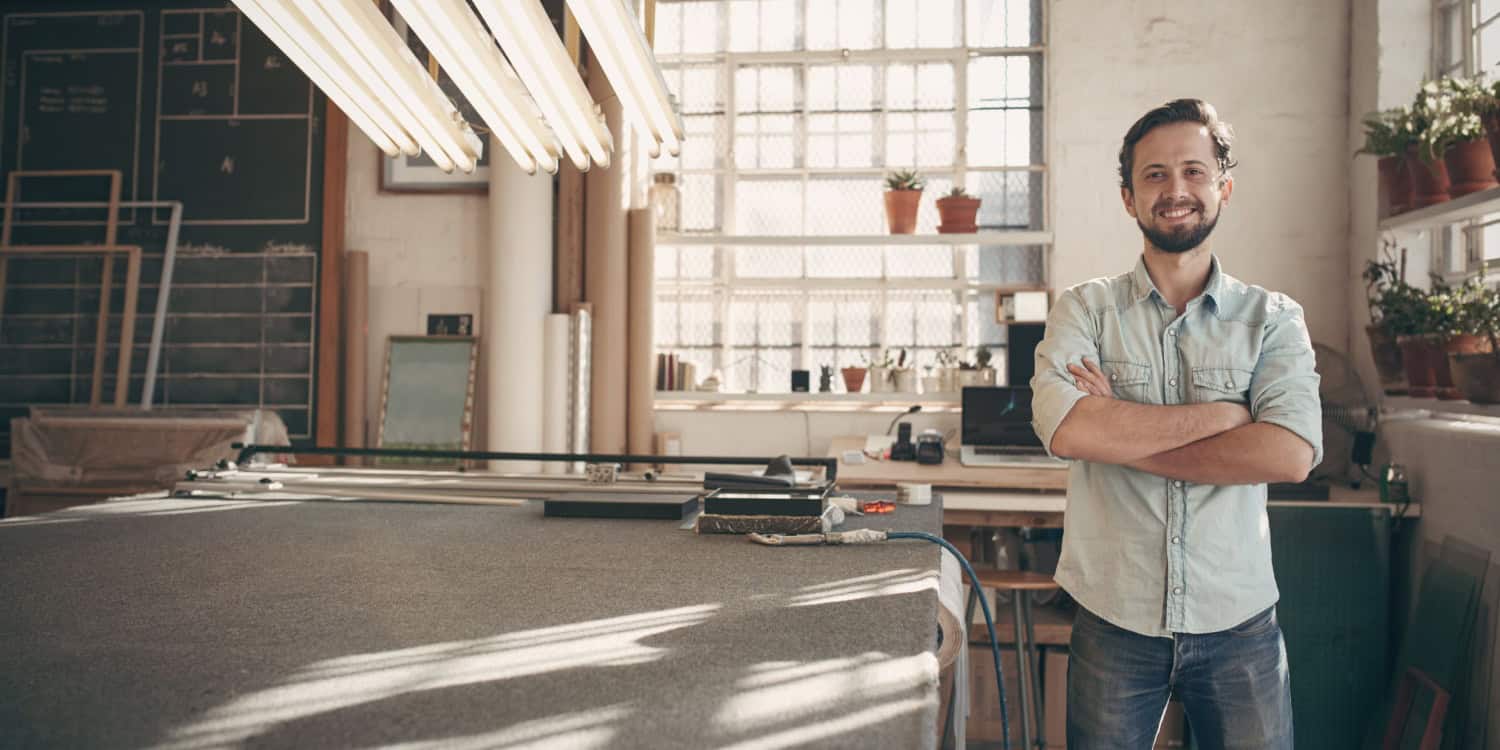 A person stands confidently in a bright workshop filled with plants and art supplies. Large windows allow natural light to illuminate the space.