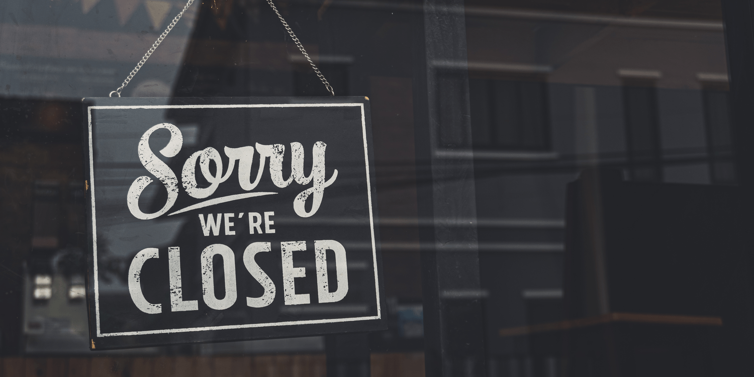 Black sign with white lettering hanging in a shop window displaying the words 'Sorry we're CLOSED'.