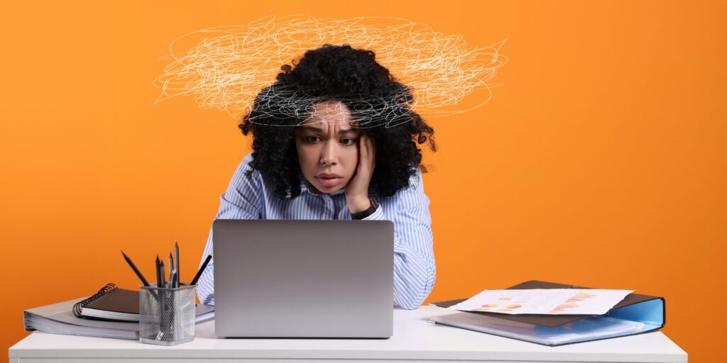 Woman looking at a laptop with a scribbled thought cloud above their head, orange background and scattered papers on the desk.