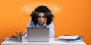 Woman looking at a laptop with a scribbled thought cloud above their head, orange background and scattered papers on the desk.