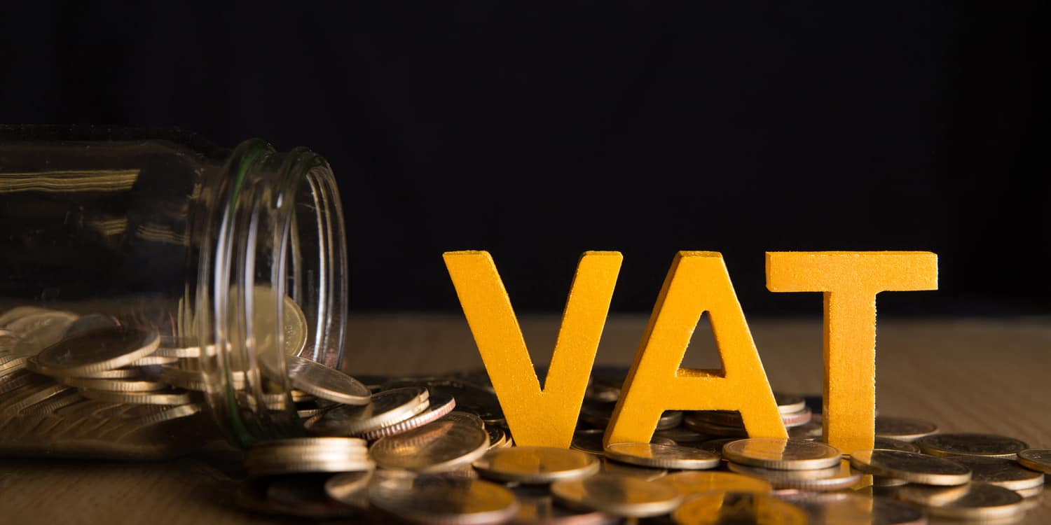 The word VAT sitting on coins and glass bottle with coins inside on black background.