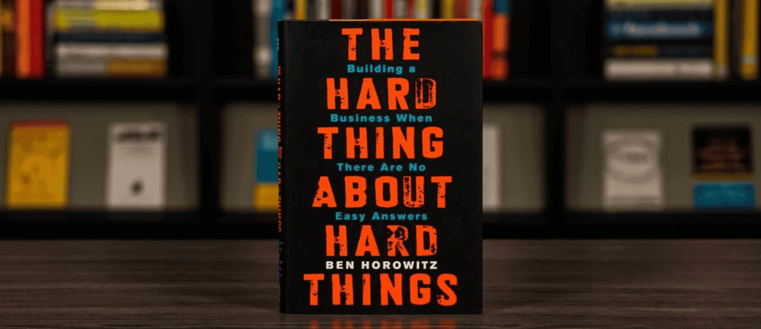 A book titled "The Hard Thing About Hard Things" by Ben Horowitz, featuring bold orange text on a black cover, displayed in front of a bookshelf.