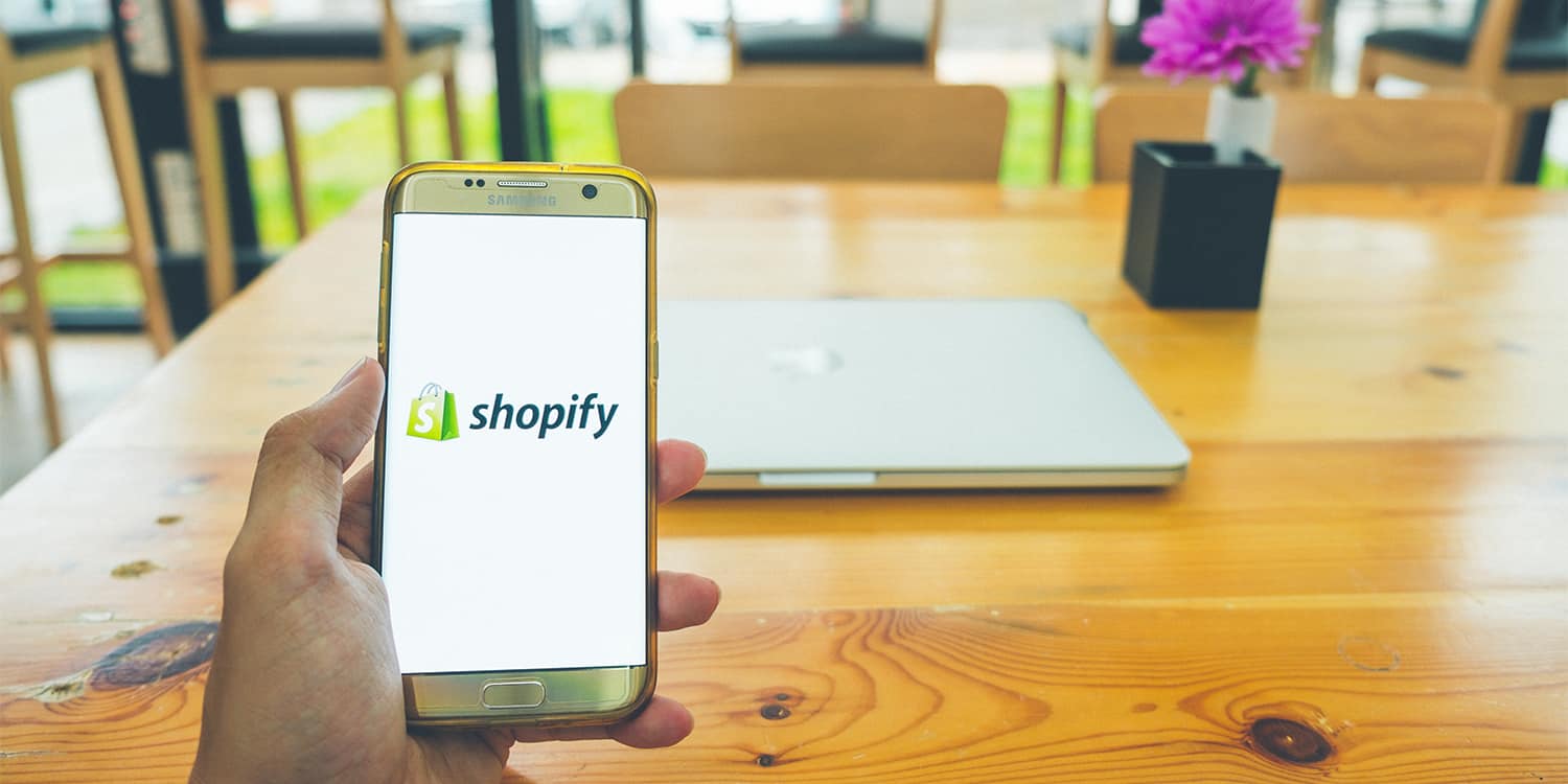 Male hand holding a smartphone with the Shopify logo displayed on the screen at an empty meeting table, illustrating the concept of the purge on meetings.