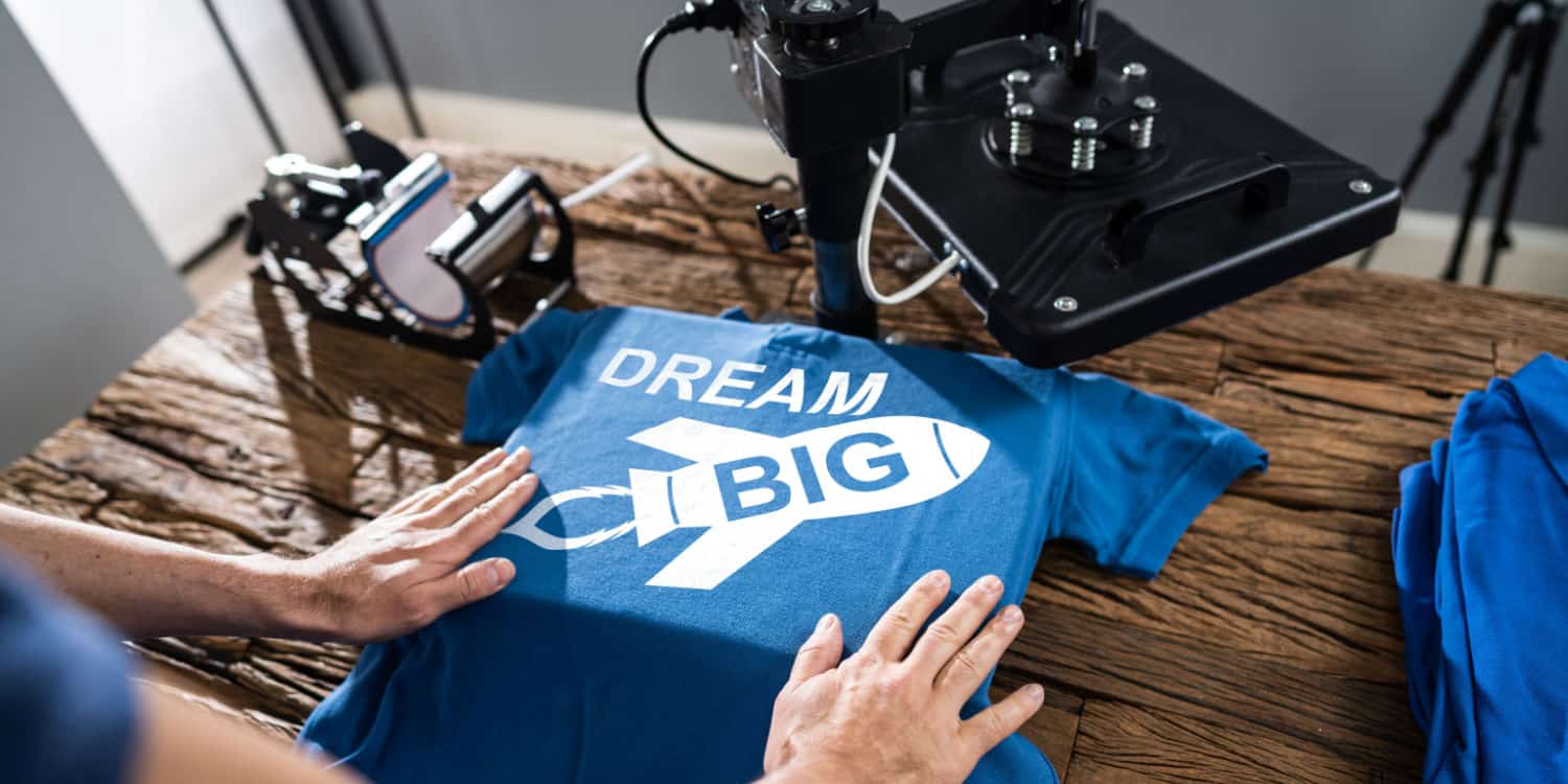 Man in a workshop printing t-shirt with rocket logo and text DREAM BIG, illustrating Print on Demand as one of the top 10 businesses to start in 2023.