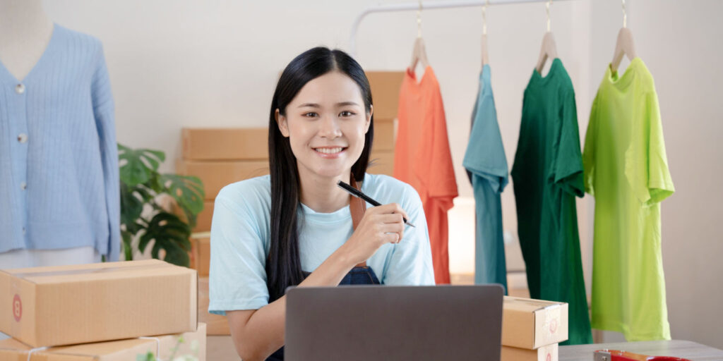 Young woman running a business from home, sitting at desk surrounded by cardboard boxes, t-shirts on garment rail and tailor's dummy.
