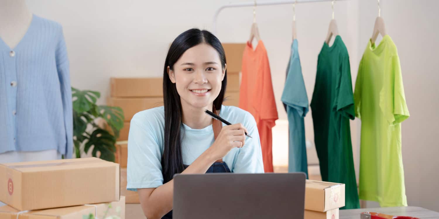 Young woman running a business from home, sitting at desk surrounded by cardboard boxes, t-shirts on garment rail and tailor's dummy.