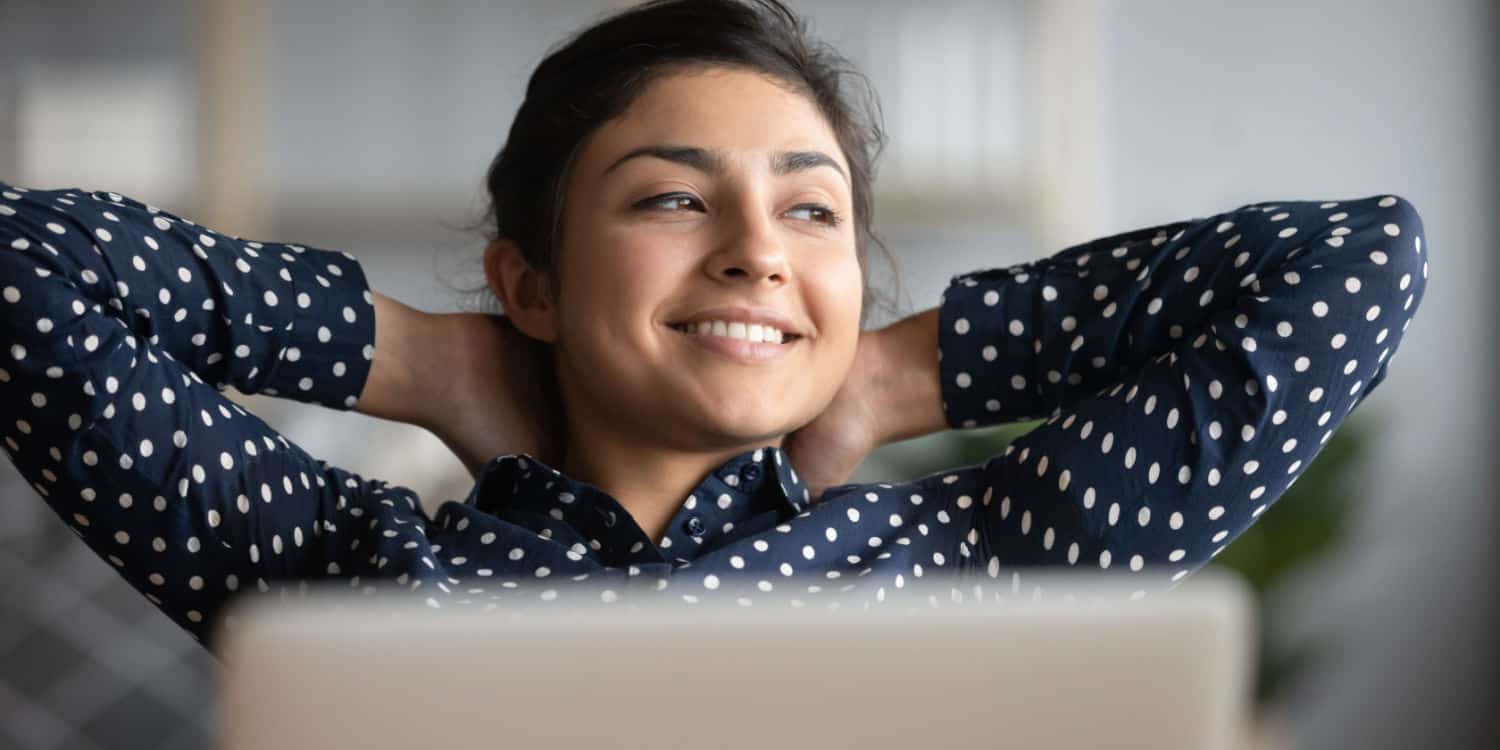 Smiling lady sat behind laptop with her arms resting behind her head, to signify job satisfaction