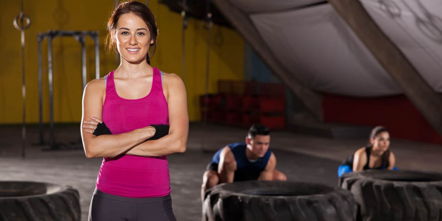 Female personal trainer standing in a gym wearing a pink sleeveless top with clients working out in the background.