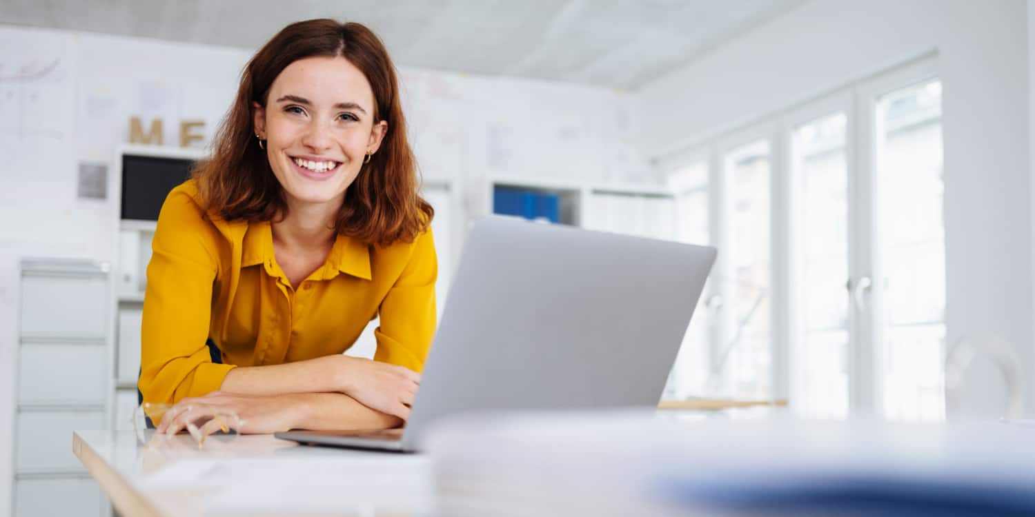 Portrait of young female personal assistant, standing at a bench desk, smiling with laptop and wearing a mustard top.