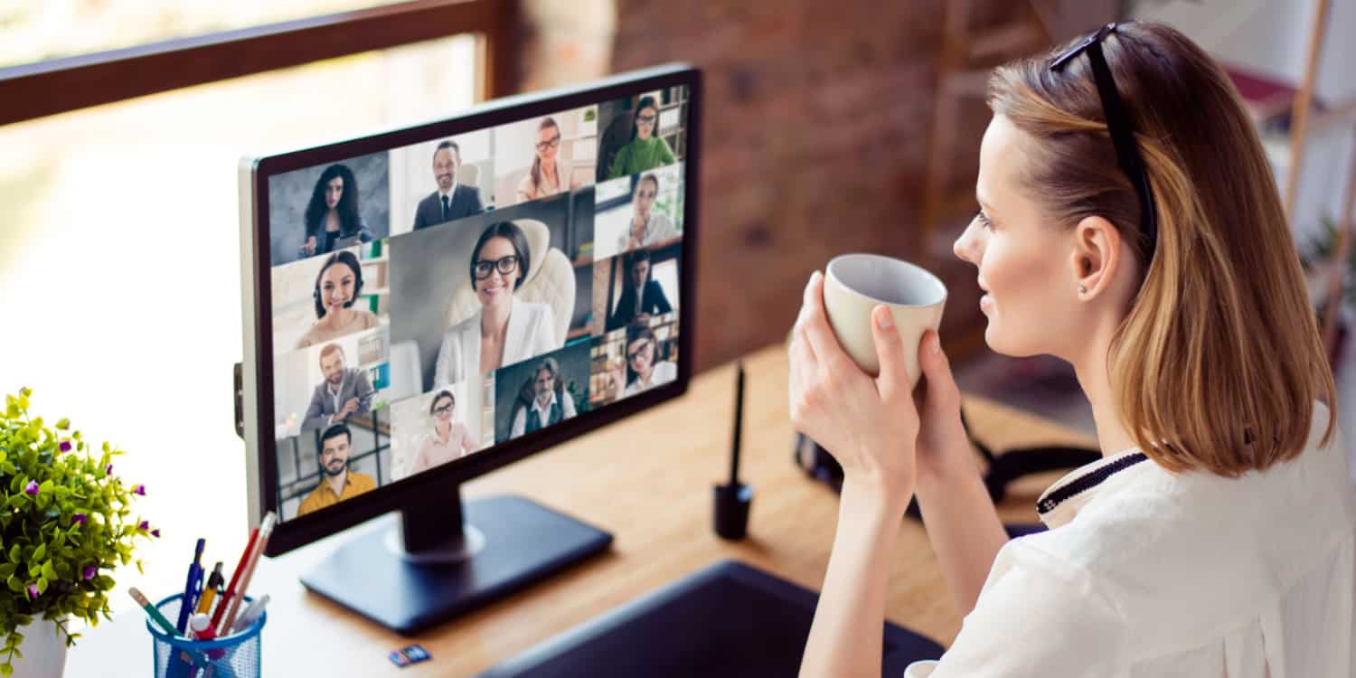 Back view photo of a female remote worker, holding a mug of coffee in two hands, and taking part in a video conference call with her coworkers.