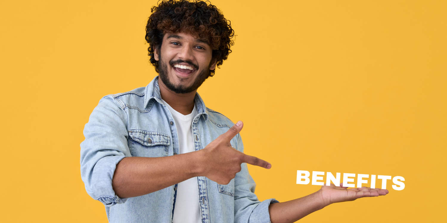 Smiling young male business owner holding a BENEFITS sign on the palm of his hand, with the other hand pointing at the sign.