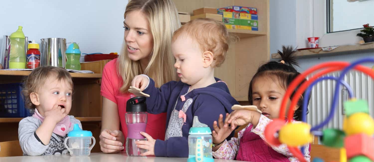 Some nursery kids on a table drinking and playing with childcare worker.