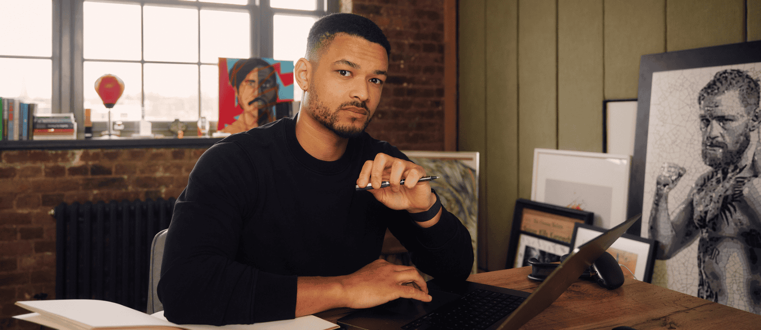Investor and entrepreneur, Steven Bartlett, sits at a desk with a laptop, holding a pen, surrounded by books, art, and large windows letting in natural light.