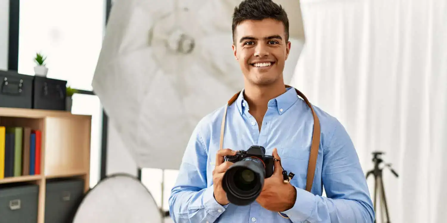 Smiling photographer holding a camera in a bright studio, with lighting gear and tripod in background.