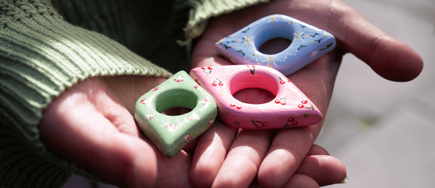 A hand holds three coluorful, hand-painted ceramic rings: green with flowers, pink with cherries, and blue with daisies.