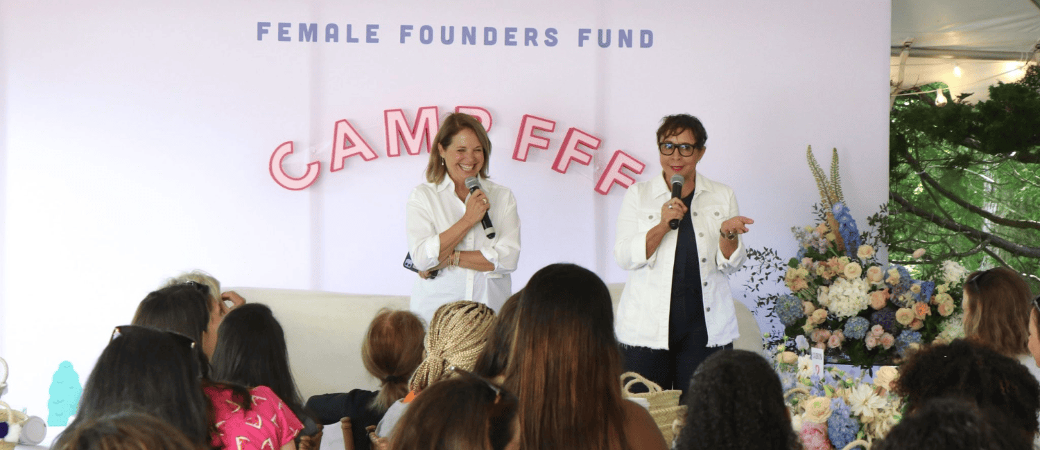 Two women from the Female Founds Fund standing with microphones in their hands, presenting a webinar to a seated audience. A sign on the wall behind them reads 'Female Founders Fund' and 'Camp FFF' below it.