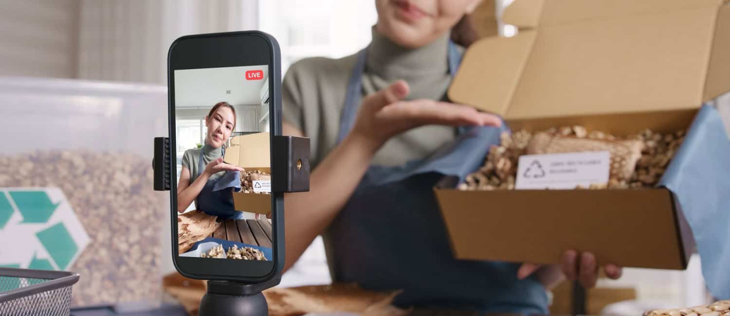 A person is live streaming on TikTok while showcasing a box filled with eco-friendly packaging materials in a bright, modern workspace.