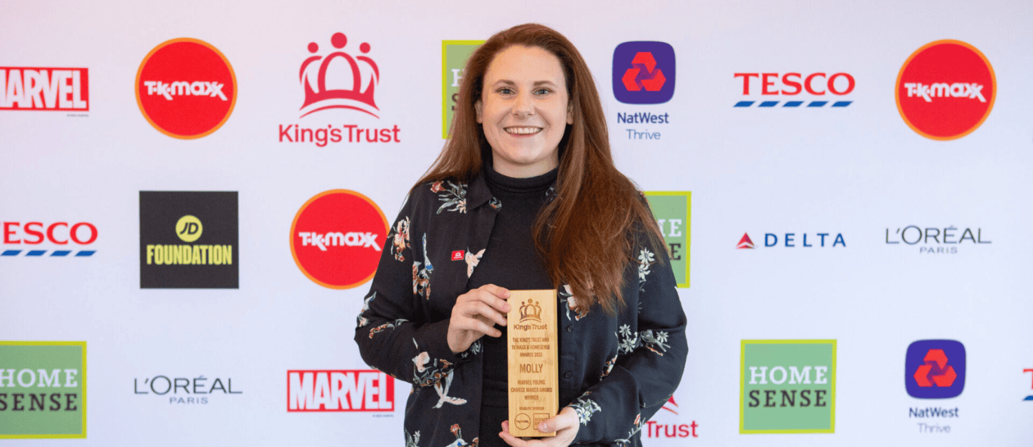 A woman smiling and holding an award in front of a sponsor backdrop featuring logos from brands like Marvel, Tesco, and L'Oréal Paris.