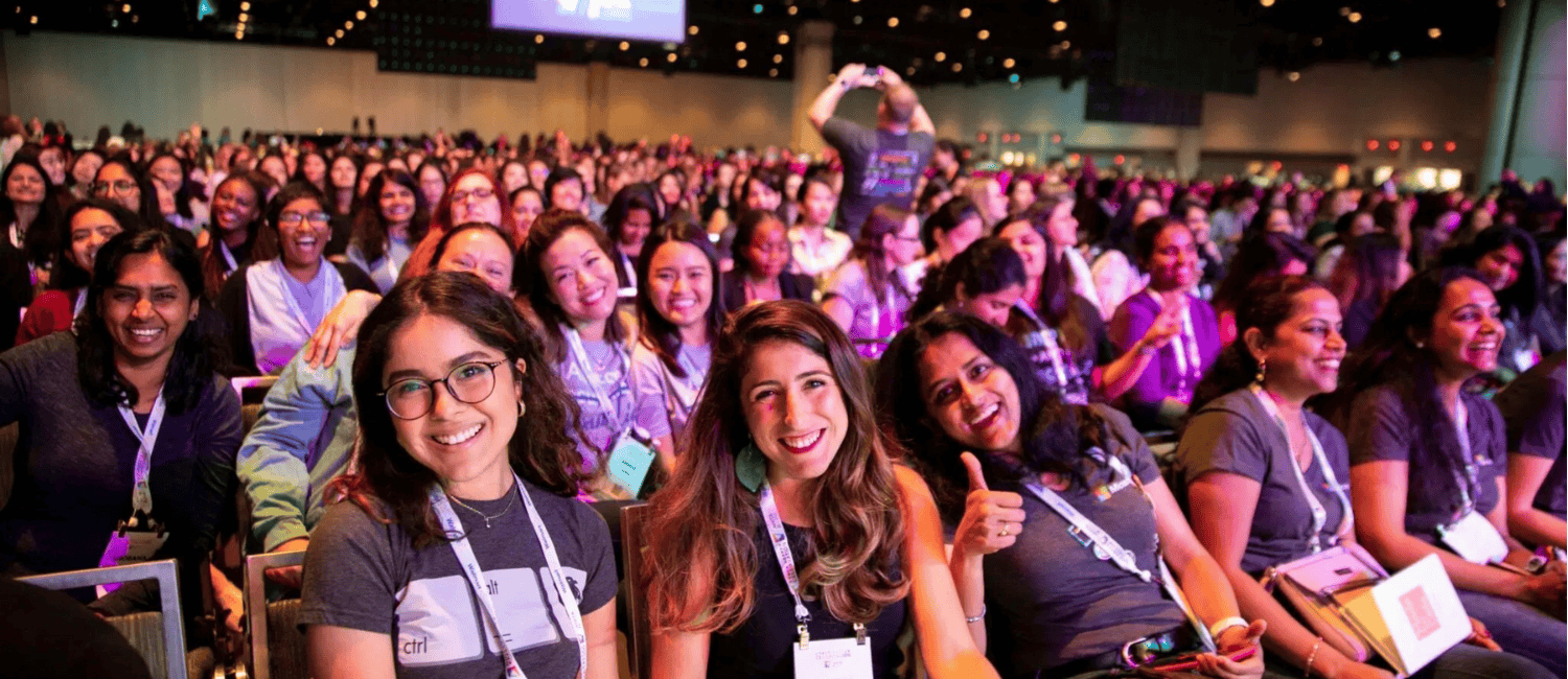 A packed auditorium filled with attendees wearing matching shirts, sitting in rows, engaged in a lively event with colourful lighting. Three happy women can be seen in the front row, smiling at the camera.