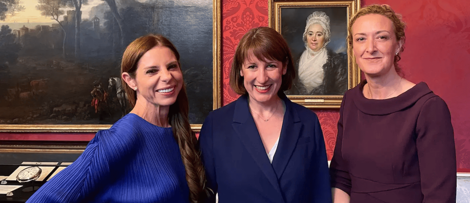 Three women stand together in a historic room adorned with red wallpaper, featuring a large landscape painting in the background.