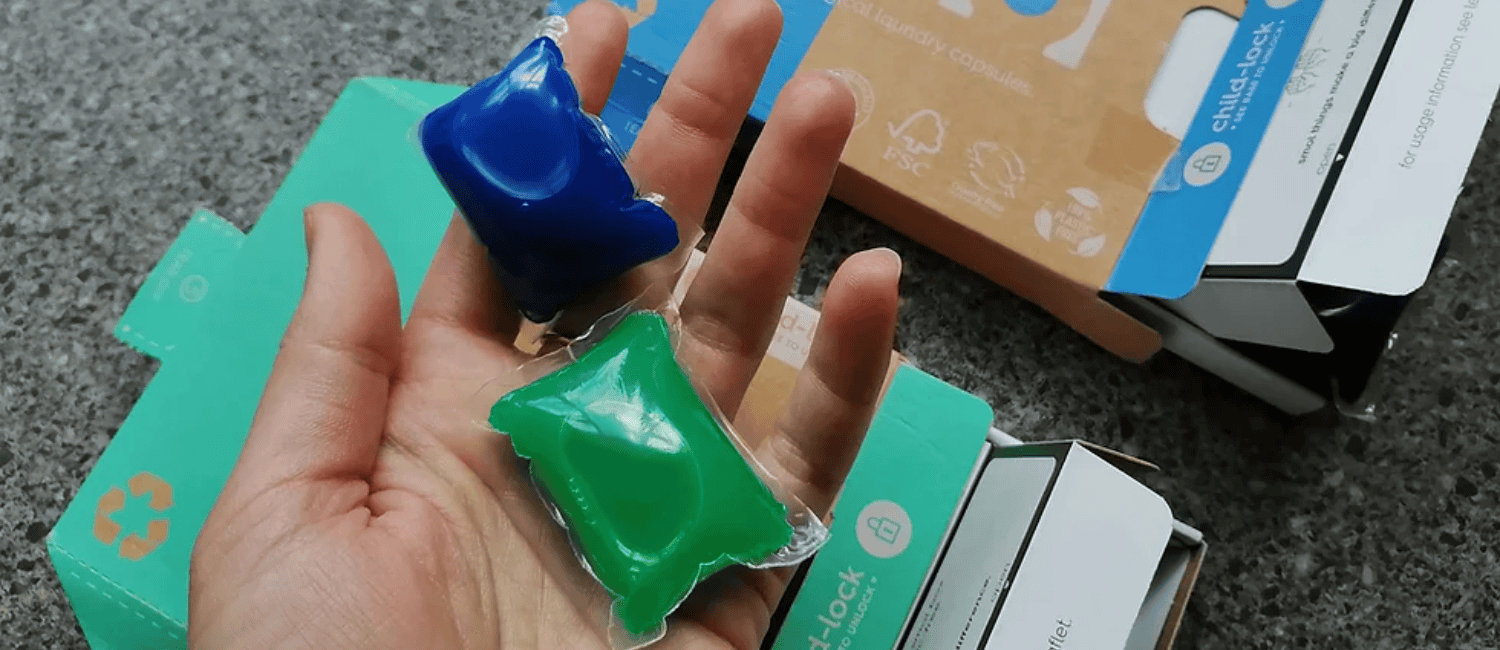 An open hand holding one blue and one green laundry capsule in its palm. One blue and one green small cardboard box can be seen below the hand.