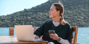 A person sits at a wooden table by the water, using a laptop while holding a smartphone, with a scenic mountainous background.