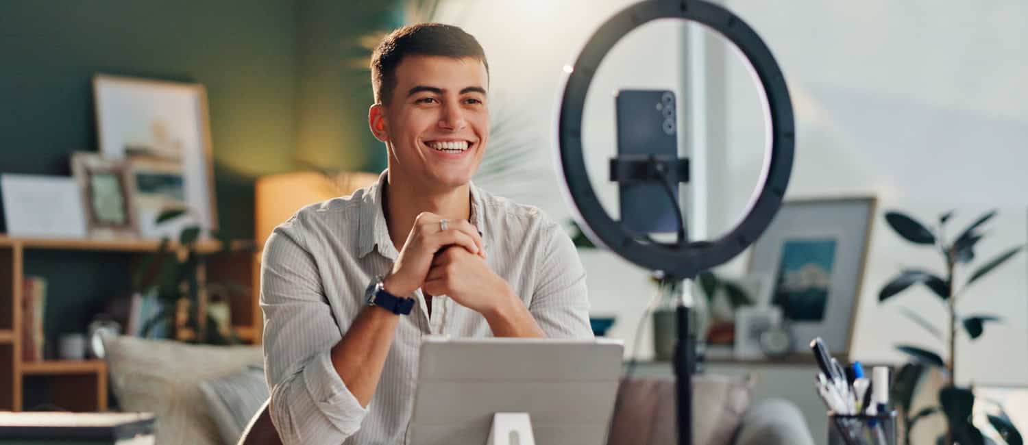 A person sits at a desk with a tablet, in front of a ring light, surrounded by plants and framed pictures in a cosy room.