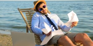 A man in a straw hat, sunglasses, shirt, tie, and shorts, sat on a sun lounger on a beach by the water. He balances a mobile phone between his head and shoulder while holding documents in each hand with a laptop next to him.