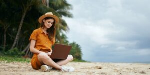 A woman in a yellow dress sitting on the beach with her laptop.