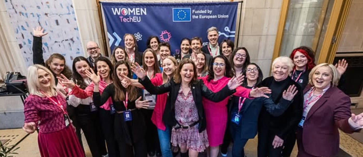Group photo of diverse women and men celebrating at the "WOMEN TechEU" event, posing in front of a banner with the European Union logo. Many attendees wear pink outfits and show enthusiastic expressions.