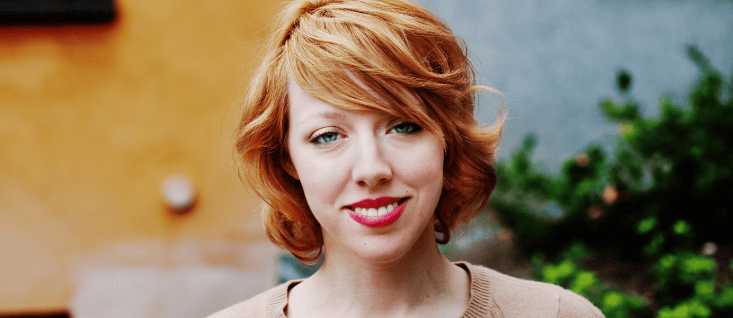 A woman with short, wavy, auburn hair stands against a blurred outdoor background of greenery and warm-toned wall, looking straight at the camera and smiling.