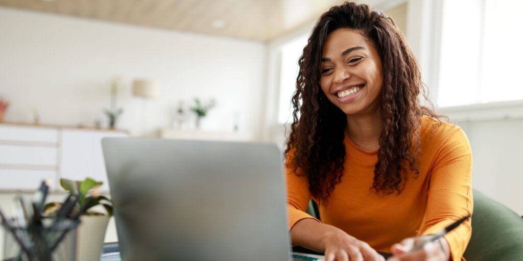 Smiling woman in an orange top watching a laptop screen and taking notes, representing someone learning from YouTube channels for entrepreneurs in a modern home office setting