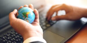 Close-up of a person holding a small globe in one hand and typing on a laptop with the other, symbolising efforts to market a business internationally and expand global reach