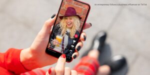 A hand holds a smartphone displaying a TikTok video of an influencer with a drink, set against a brick background.