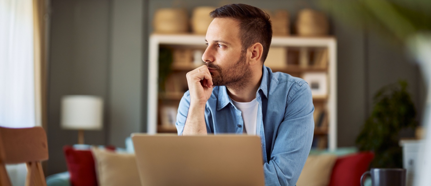 Entrepreneur sitting at a desk with a laptop, resting his chin on his hand and looking thoughtfully into the distance in a cosy home office setting