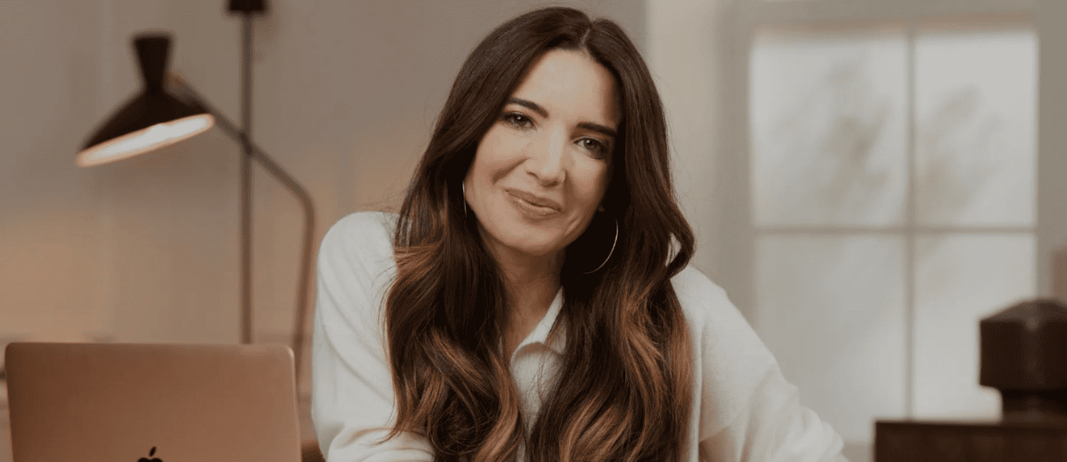 A woman with long brown hair and hoop earrings smiles warmly, sitting in a cosy room with a laptop. Soft lighting and a lamp create a calm atmosphere.