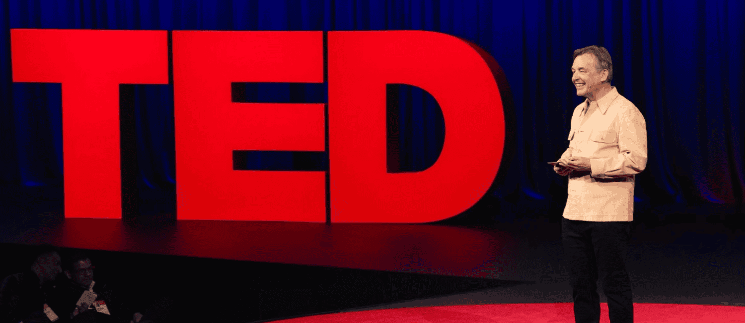 A speaker in a beige shirt smiles on a TED stage, holding notes. Large red "TED" letters are behind him. The atmosphere is engaging and professional.