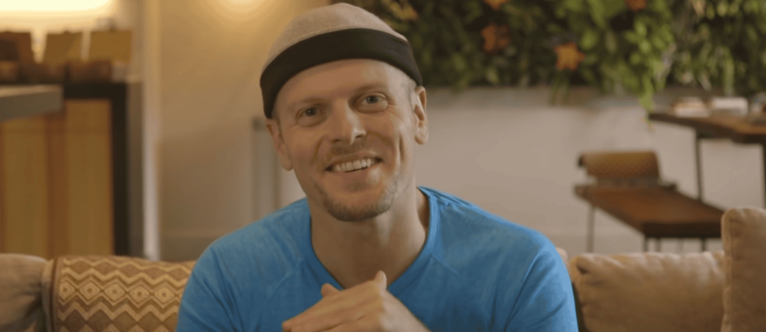 Smiling man with a headband in a blue shirt sits on a couch in a dimly-lit room with plants, with one hand placed inside the other