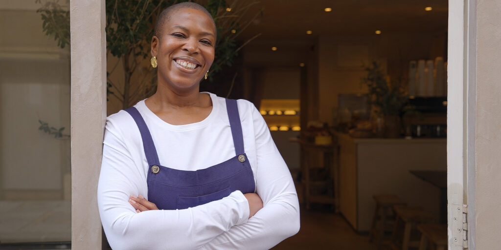 Smiling entrepreneur standing confidently in a doorway, arms crossed, wearing a white shirt and navy apron, with a warmly lit café interior in the background