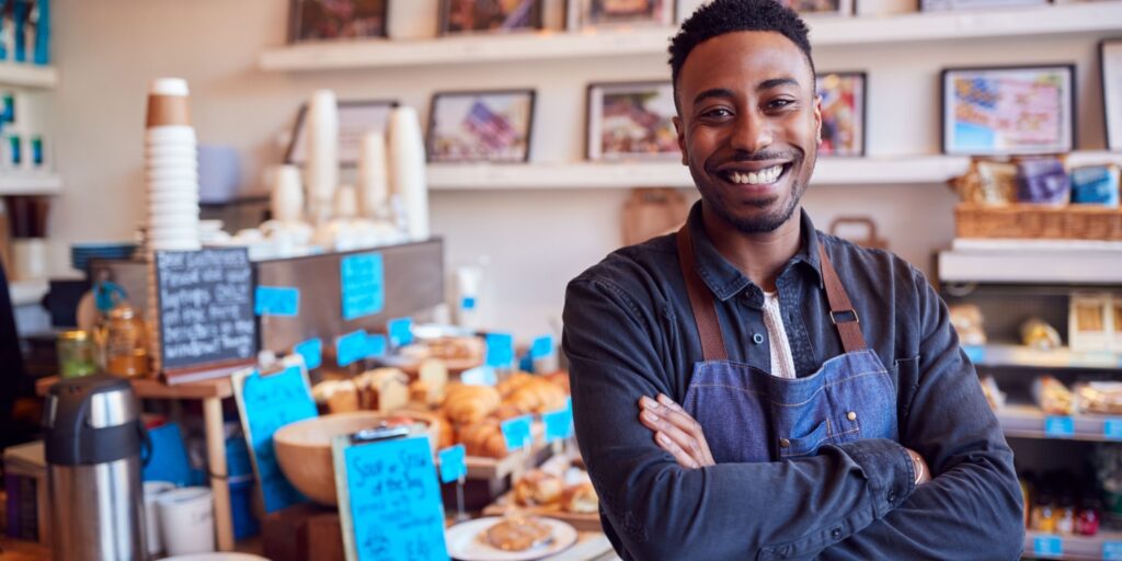 A barista in a denim apron stands confidently, arms crossed and smiling, in a café filled with baked goods and colourful signage.