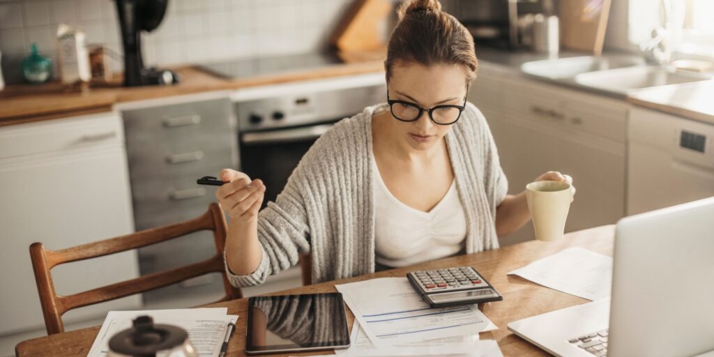 Business owner reviewing financial paperwork at home while working on a laptop, calculating figures with a pen and calculator in hand, preparing to file a CT600 Corporation Tax Return for their UK limited company