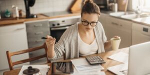 Business owner reviewing financial paperwork at home while working on a laptop, calculating figures with a pen and calculator in hand, preparing to file a CT600 Corporation Tax Return for their UK limited company