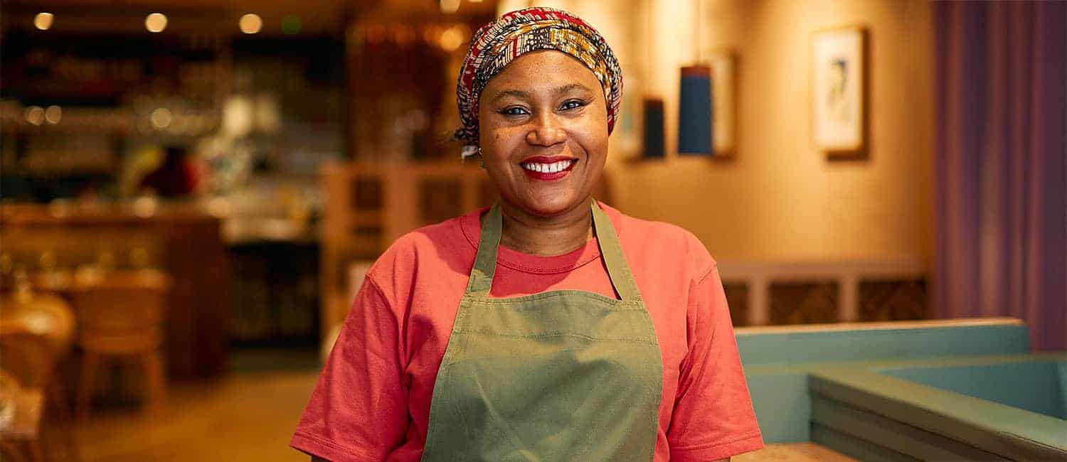 Adejoké Bakare, founder of Chishuru, a Michelin-starred Black-owned West African restaurant, smiles warmly while wearing an apron inside her vibrant dining space