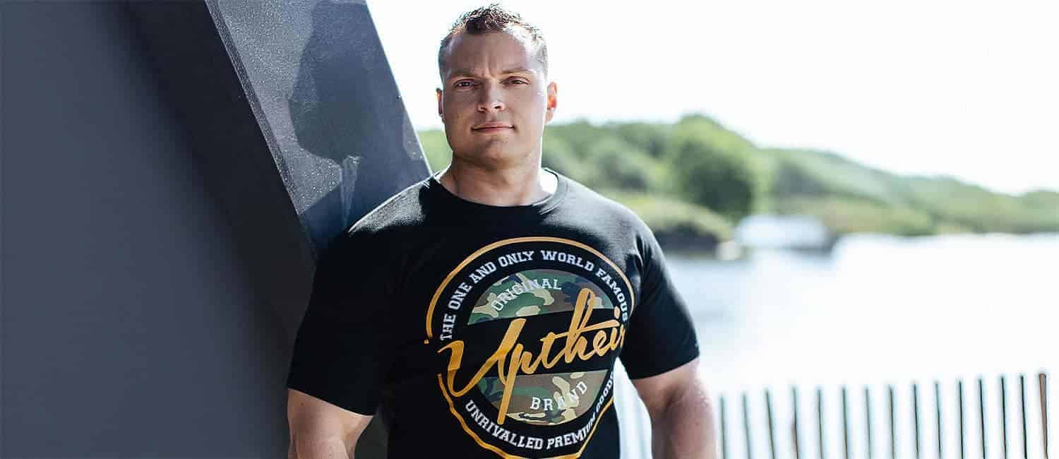 Ben Pearson, Founder of BigClothing4U and Uptheir, stands confidently outdoors in the sunlight next to a modern structure, with a lake and greenery in the background. He wears a black t-shirt featuring a bold circular "Uptheir Brand" logo with a camouflage design