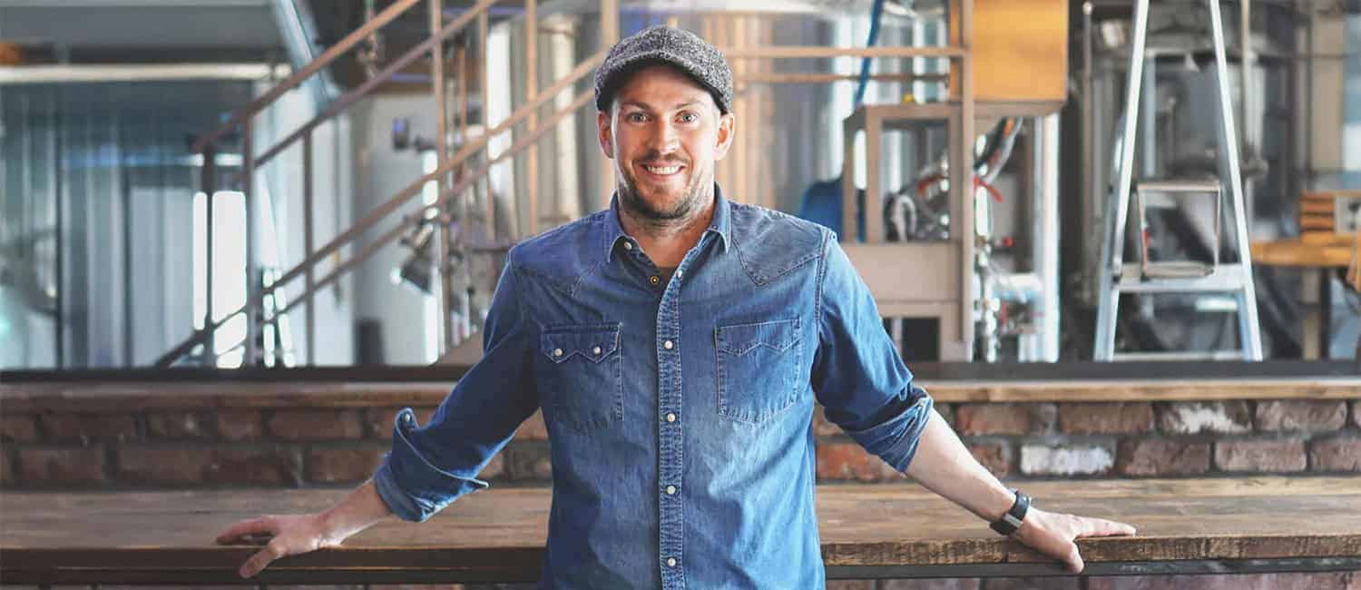 James Watt, Co-founder of BrewDog, wearing a denim shirt and a grey flat cap smiles while leaning on a wooden bar counter in an industrial-style brewery setting with stainless steel brewing equipment in the background