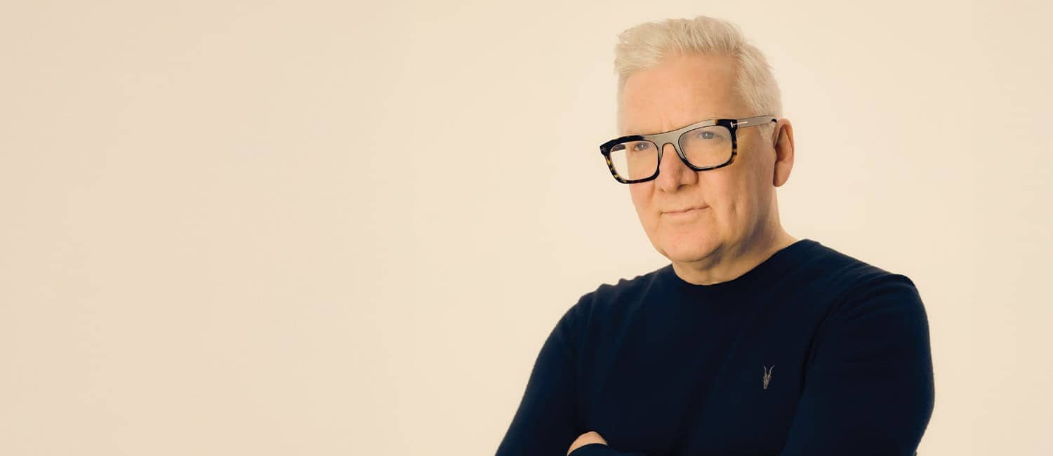 A person with slicked-back gray hair and glasses stands confidently with arms crossed, wearing a dark top against a light background.